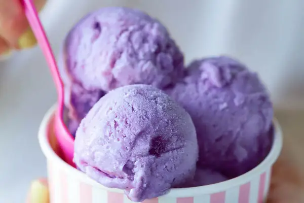 Tre palline di gelato viola in una coppetta a righe rosa e bianche, con un cucchiaino rosa. Un dolce piacere che non causa stitichezza.